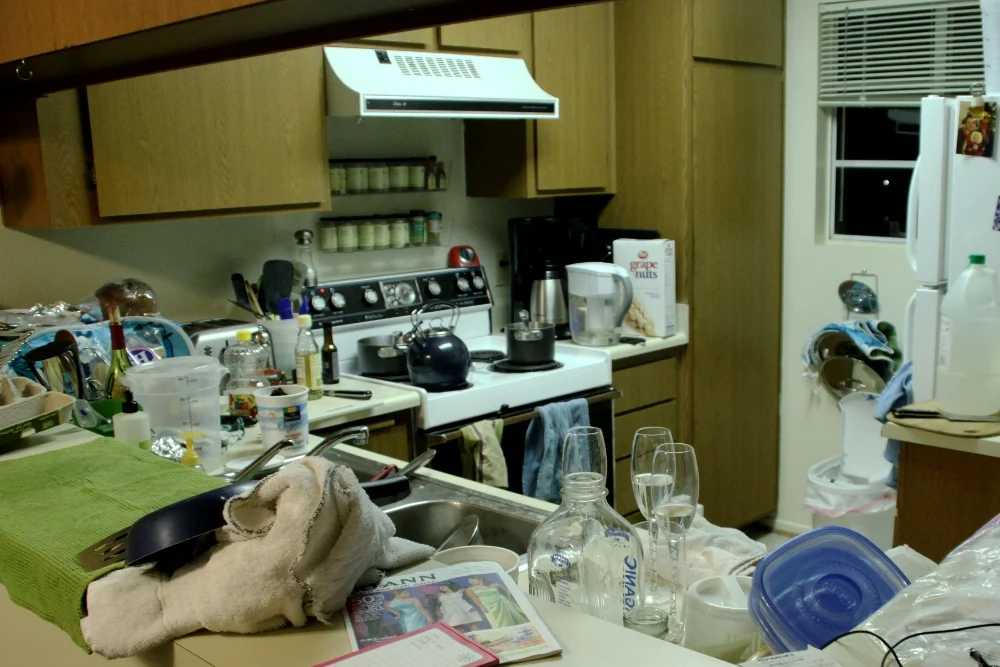 A real home kitchen before cleanup, with dishes, towels, and containers cluttering the counters